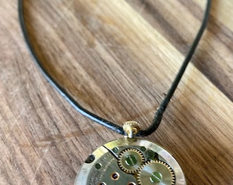 May include: A vintage watch movement on a black leather cord necklace. The watch face is exposed, showing the intricate gears and mechanics.