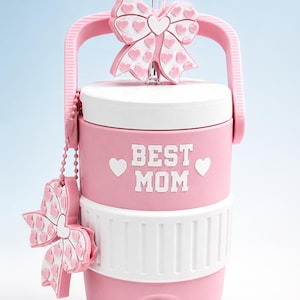 May include: Pink water cooler with a white lid and base, featuring the text "BEST MOM" in white. A pink bow with heart accents adorns the handle and a smaller bow hangs from a chain. Includes a clear straw.