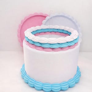 May include: A white cylindrical cake dummy featuring a blue scalloped border at the base. The top is decorated with three stacked scalloped rings in white, blue, and pink. Two additional scalloped circular discs in pink and light blue stand behind.