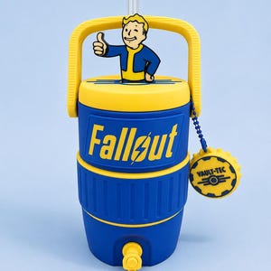 May include: A blue and yellow beverage cooler featuring the Fallout logo and a Vault Boy figure on the lid. The cooler has a yellow handle, a clear straw, and a blue beaded chain with a circular Vault-Tec tag attached to the side of the lid.