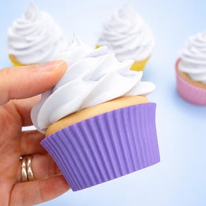May include: A cupcake with a light tan base, topped with a swirl of white frosting, and encased in a ribbed purple silicone liner. Additional cupcakes with white frosting and yellow or pink liners are visible in the background.