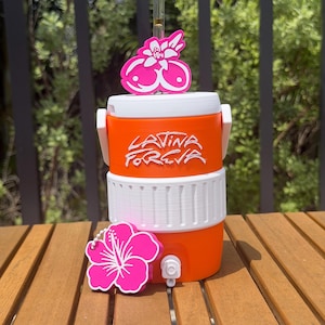 May include: An orange and white beverage cooler featuring the text "LATINA FOREVA" in white script. The cooler includes a clear straw with a pink floral topper and a separate pink hibiscus flower charm attached to the front near the white spigot.
