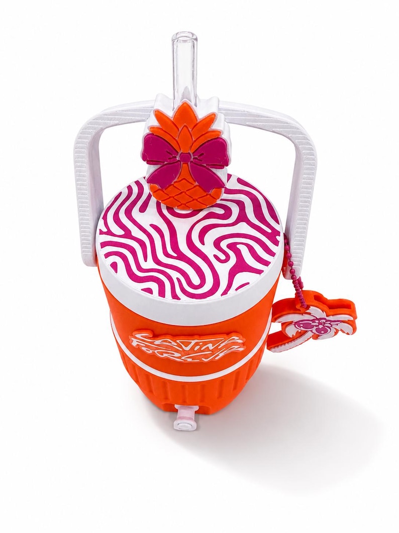 May include: An orange beverage cooler with a white handle and a white lid featuring a pink wavy pattern. The lid has a pineapple topper with a pink bow. A clear straw protrudes from the top. The cooler features the text "Living For It" and a palm tree charm.