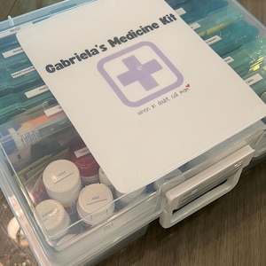 May include: A clear plastic medicine box with a white label that says "Gabriela's Medicine Kit" with a purple cross and the text "when in doubt, call mom". The box is filled with various medications and supplies.