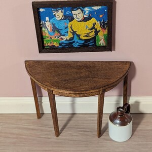 May include: Miniature wooden half-circle table with four legs. A framed Star Trek art print hangs above the table. A small brown and white jug sits on the floor next to the table. The table and jug are made of wood and ceramic.