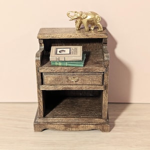May include: A miniature wooden nightstand with a drawer and open shelf. A small gold elephant figurine sits on top, with two miniature books on the middle shelf. The nightstand has a distressed, aged appearance.