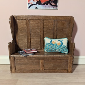 May include: A miniature wooden bench with a high back and armrests. A small, embroidered pillow with two peacocks in teal and orange sits on the bench. Two small books are on the bench. The bench is a dark brown color.