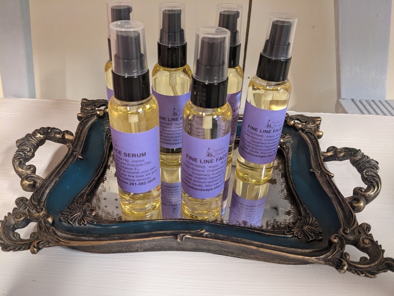 K&ouml;nnte beinhalten: Sechs kleine Glasflaschen mit Tropfern, gef&uuml;llt mit einer klaren Fl&uuml;ssigkeit. Die Flaschen sind mit "Fine Line Face Serum" und "Face Serum" beschriftet, mit einem violetten Hintergrund und wei&szlig;em Text. Die Flaschen sind auf einem dekorativen, verspiegelten Tablett mit goldenen Akzenten angeordnet.