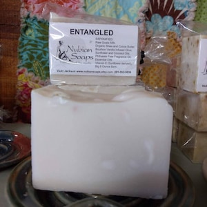 May include: A white bar of soap labeled "Entangled" with the text "Nubian Soaps" and ingredients listed. The soap is on a blue and brown ceramic dish.