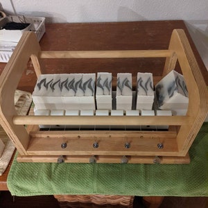 May include: A wooden soap cutting frame with a set of white and gray swirl patterned soap bars. The frame has a wooden handle and a metal wire grid.
