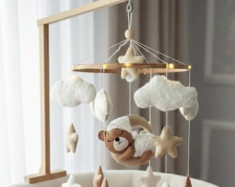 May include: A wooden baby mobile with a light brown frame. The mobile features white clouds, beige stars, and a sleeping teddy bear on a crescent moon. The mobile is suspended above a white crib.