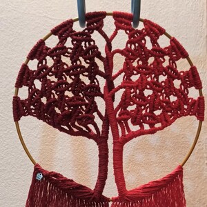 May include: A red macrame wall hanging in the shape of a tree with a gold hoop and fringe.