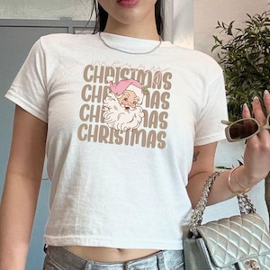 以下が含まれることがあります： サンタクロースの茶色のグラフィックと「メリークリスマス」というテキストが積み重ねられたパターンで繰り返されている白いクロップドTシャツです。