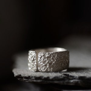 May include: A silver ring with a textured surface. The ring is wide and has a square shape.