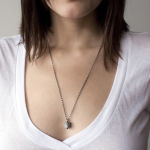 May include: A silver chain necklace with a small, cylindrical pendant. The pendant has a textured surface and a small, clear stone in the center.