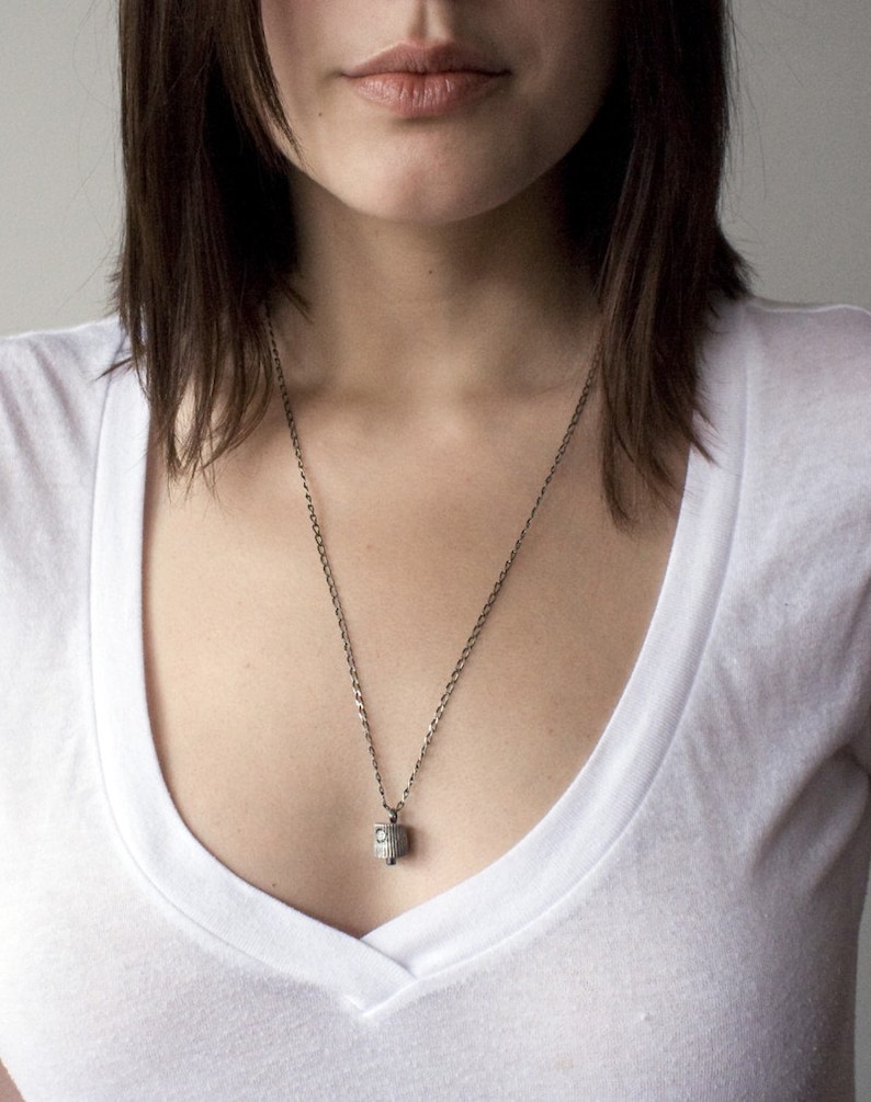 May include: A silver chain necklace with a small, cylindrical pendant. The pendant has a textured surface and a small, clear stone in the center.
