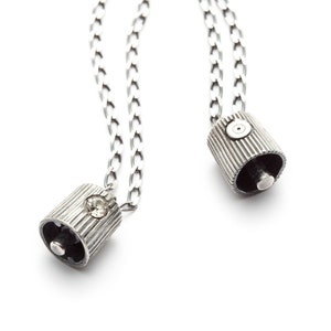 May include: Two silver chain necklaces with silver pendant charms. One charm is a small cylinder with a single rhinestone, the other charm is a cylinder with a small circle on the top.