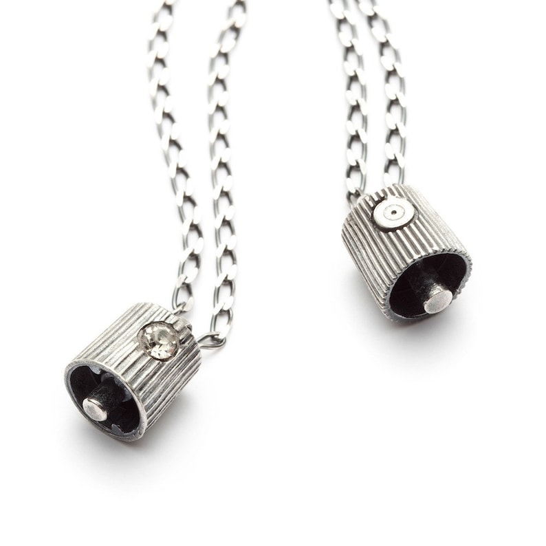 May include: Two silver chain necklaces with silver pendant charms. One charm is a small cylinder with a single rhinestone, the other charm is a cylinder with a small circle on the top.