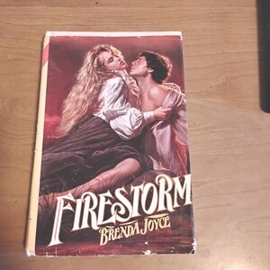 May include: A paperback book titled "Firestorm" by Brenda Joyce. The cover art depicts a romantic scene with a woman in a white blouse and a man in a pink garment. The background is a deep red.