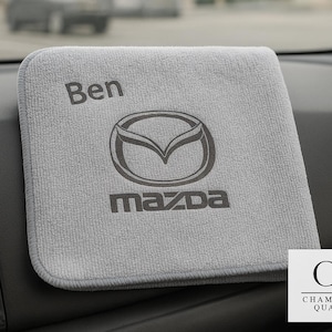 May include: A gray microfiber cleaning cloth with the Mazda logo and the name "Ben" printed on it. The cloth is square and sits on a car dashboard. The Mazda logo is in the center, with the brand name below it.