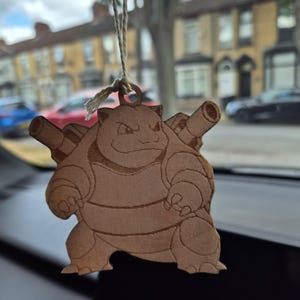 May include: A light brown wooden hanging ornament shaped like the character Blastoise, featuring engraved details of the creature with two cannons on its back. The ornament is suspended by a thin, light-colored string attached to a small circular loop.