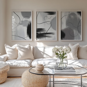 Könnte beinhalten: Drei gerahmte abstrakte Kunstdrucke in Grau- und Schwarztönen hängen über einem weißen Sofa mit cremefarbenen Kissen. Ein Glastisch und gewebte Ottomanen vervollständigen die Wohnzimmerdekoration.