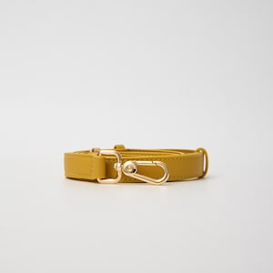 K&ouml;nnte beinhalten: Ein senfgelber Taschenriemen mit glatter, matter Oberfl&auml;che und goldfarbenen Metallbeschl&auml;gen. Der Riemen verf&uuml;gt &uuml;ber einen drehbaren Karabinerhaken zur Befestigung an einer Tasche sowie eine rechteckige Schnalle zur L&auml;ngenverstellung.