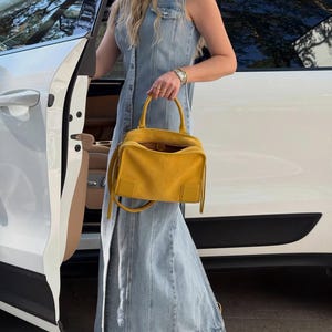 K&ouml;nnte beinhalten: Ein &auml;rmelloses Maxikleid aus hellblauem Denim mit durchgehender Knopfleiste, kombiniert mit einer leuchtend gelben Wildlederhandtasche mit zwei Tragegriffen und abnehmbarem Schulterriemen sowie hellgelben Riemchensandalen mit Absatz.