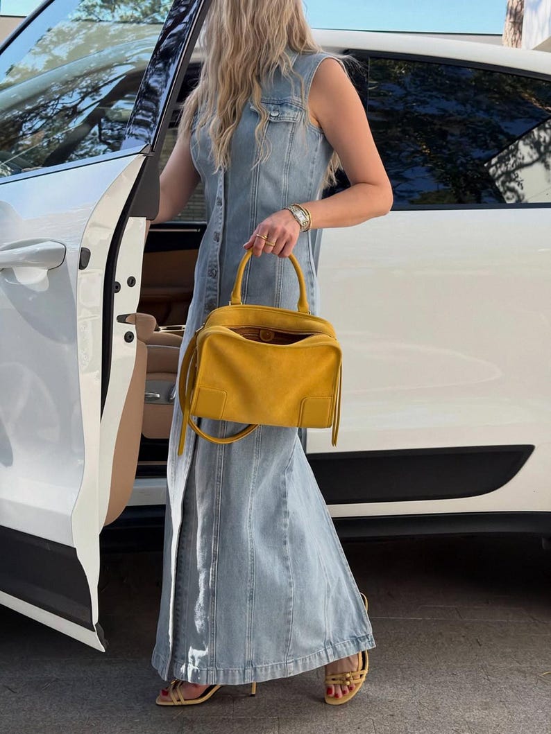 K&ouml;nnte beinhalten: Ein &auml;rmelloses Maxikleid aus hellblauem Denim mit durchgehender Knopfleiste, kombiniert mit einer leuchtend gelben Wildlederhandtasche mit zwei Tragegriffen und abnehmbarem Schulterriemen sowie hellgelben Riemchensandalen mit Absatz.