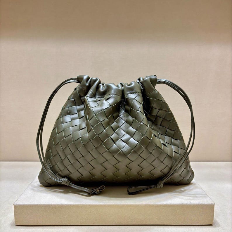 May include: An olive green leather pouch featuring a woven intrecciato pattern. The bag has a gathered top closure with thin leather drawstring cords on each side that hang down, creating a soft, rounded shape with a structured woven texture.