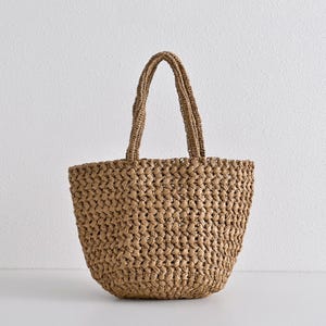 May include: A woven tote bag in a natural tan color. The bag features a sturdy, open weave design and two braided handles. The bag is designed to be carried by hand or over the shoulder. The bag is made of a natural fiber material.