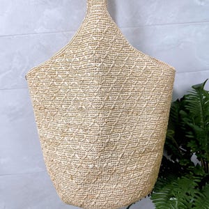 May include: A woven straw tote bag with a diamond pattern. The bag has a wide opening and a handle at the top. The bag is a natural beige color and appears to be handmade.