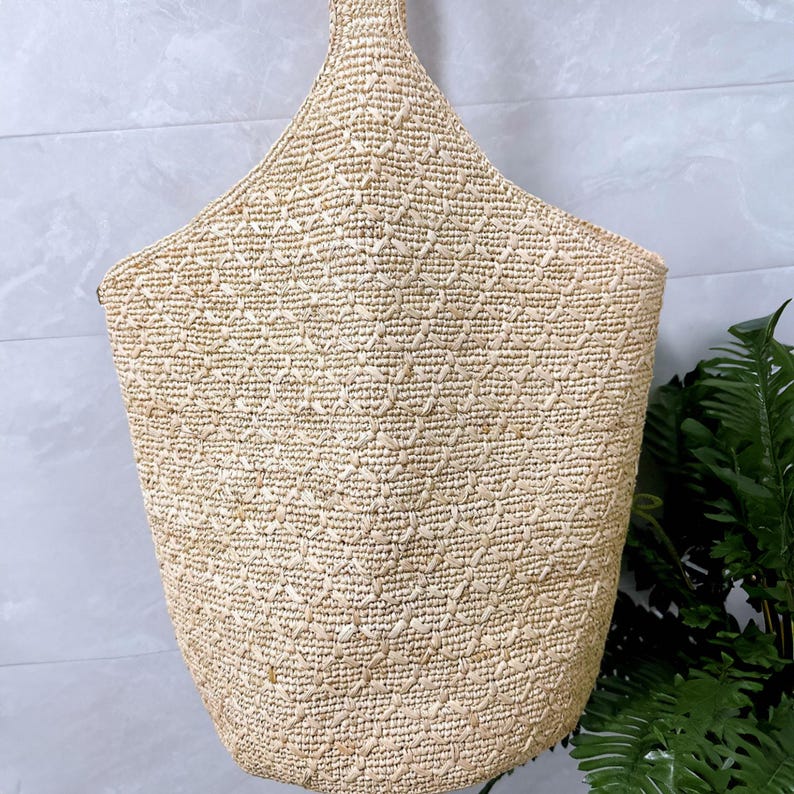May include: A woven straw tote bag with a diamond pattern. The bag has a wide opening and a handle at the top. The bag is a natural beige color and appears to be handmade.