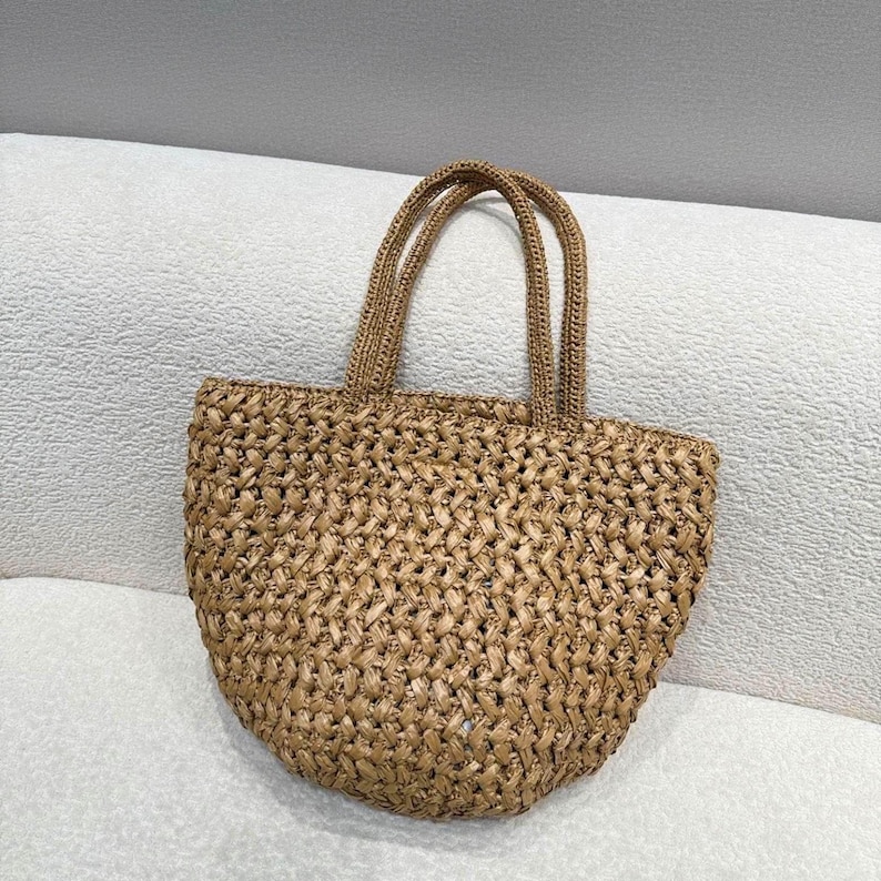 May include: A woven straw tote bag with two handles. The bag is a light brown color and has a rounded shape. The weave pattern is visible throughout the bag, creating a textured appearance. The handles are also made of woven straw.