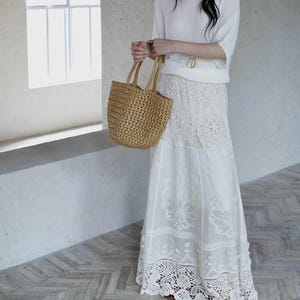May include: A woman's outfit featuring a white ribbed knit top, a long white lace skirt, and a woven straw tote bag. The skirt has intricate floral lace detailing. The outfit is paired with brown sandals.