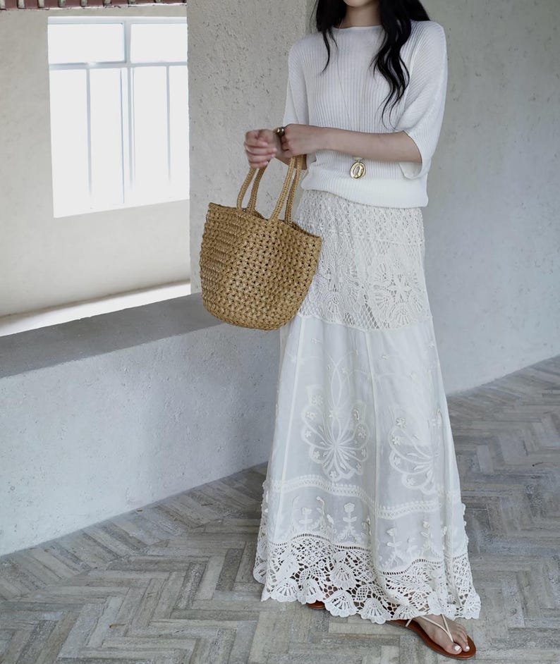 May include: A woman's outfit featuring a white ribbed knit top, a long white lace skirt, and a woven straw tote bag. The skirt has intricate floral lace detailing. The outfit is paired with brown sandals.