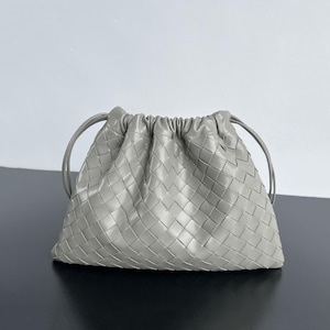 May include: A light gray pouch handbag featuring a woven leather exterior with a grid pattern. The bag has a gathered top opening with thin leather drawstrings on each side, creating a soft, rounded shape with a smooth, uniform texture.