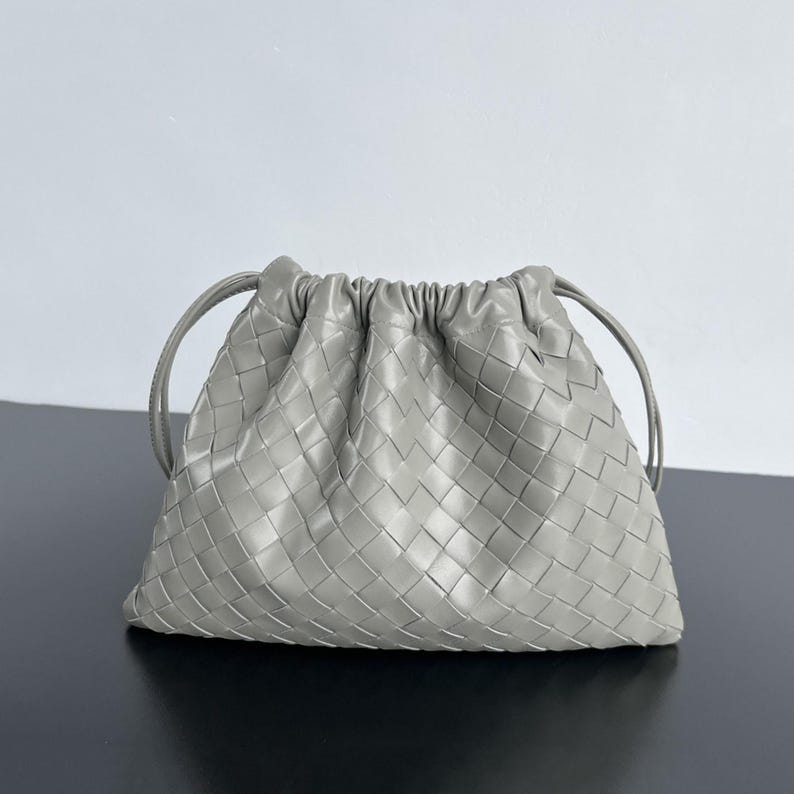 May include: A light gray pouch handbag featuring a woven leather exterior with a grid pattern. The bag has a gathered top opening with thin leather drawstrings on each side, creating a soft, rounded shape with a smooth, uniform texture.
