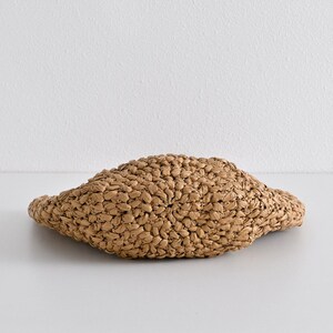 May include: A woven straw hat in a natural tan color. The hat has a rounded crown and a slightly curved brim. The texture of the straw is visible, with a tight weave pattern. The hat is sitting on a white surface.