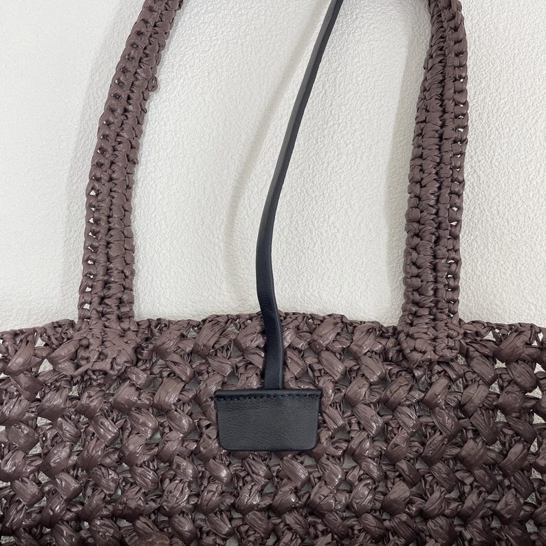 May include: A woven brown tote bag with two braided handles and a black leather strap and tag. The bag has a textured, basket-weave design. The handles are thick and sturdy, and the strap is thin and elegant.