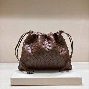May include: A dark brown leather pouch featuring a woven intrecciato pattern. The bag has a soft, slouchy silhouette with a drawstring closure at the top, secured by thin leather cords that extend from the sides to create a gathered effect.
