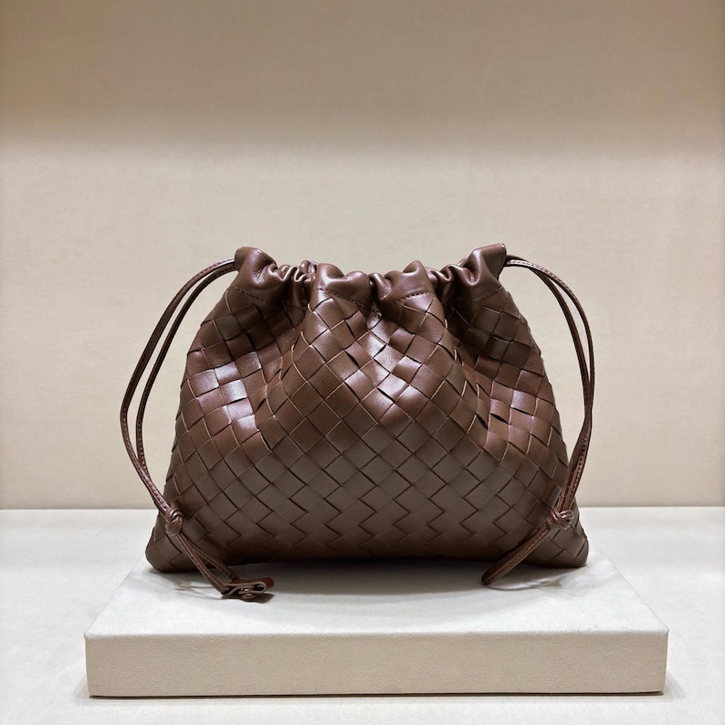 May include: A dark brown leather pouch featuring a woven intrecciato pattern. The bag has a soft, slouchy silhouette with a drawstring closure at the top, secured by thin leather cords that extend from the sides to create a gathered effect.