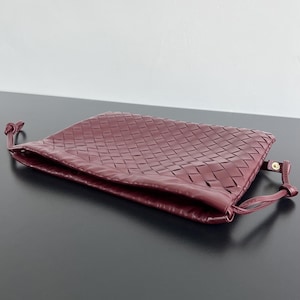May include: A deep burgundy clutch bag featuring a woven leather exterior pattern. The rectangular bag has a soft, flexible structure with thin leather side straps that include small gold-toned metal hardware accents at the attachment points.