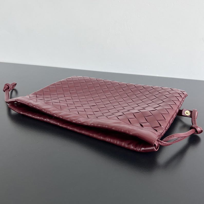 May include: A deep burgundy clutch bag featuring a woven leather exterior pattern. The rectangular bag has a soft, flexible structure with thin leather side straps that include small gold-toned metal hardware accents at the attachment points.