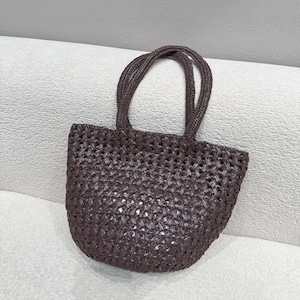 May include: A woven tote bag in a dark brown color. The bag has a rounded shape with two handles. The weave pattern is visible, creating a textured appearance. The bag is designed for carrying items.