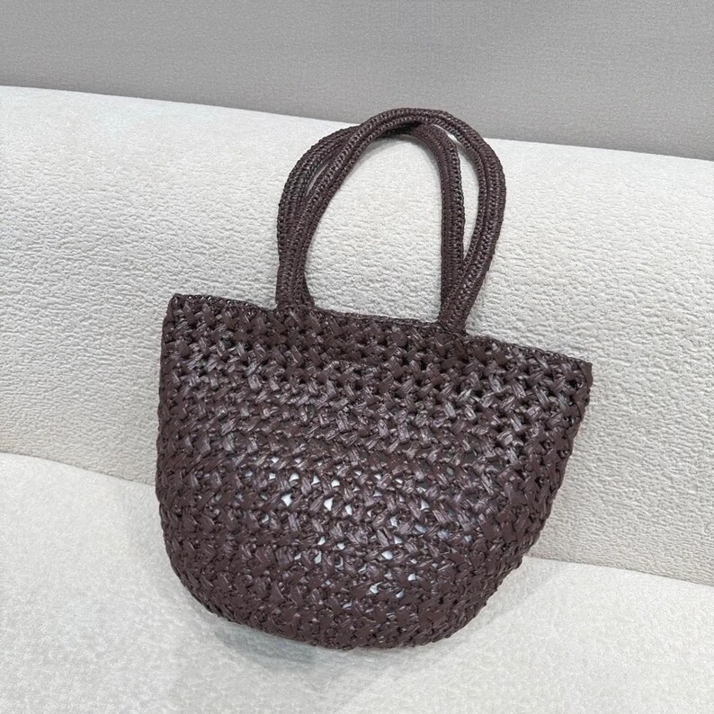May include: A woven tote bag in a dark brown color. The bag has a rounded shape with two handles. The weave pattern is visible, creating a textured appearance. The bag is designed for carrying items.