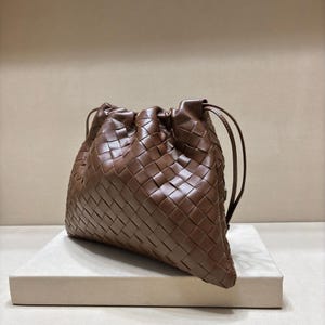 May include: A brown leather handbag featuring a woven intrecciato pattern throughout the exterior. The bag has a soft, slouchy structure with a drawstring closure at the top, secured by thin leather cords that extend from the sides of the opening.