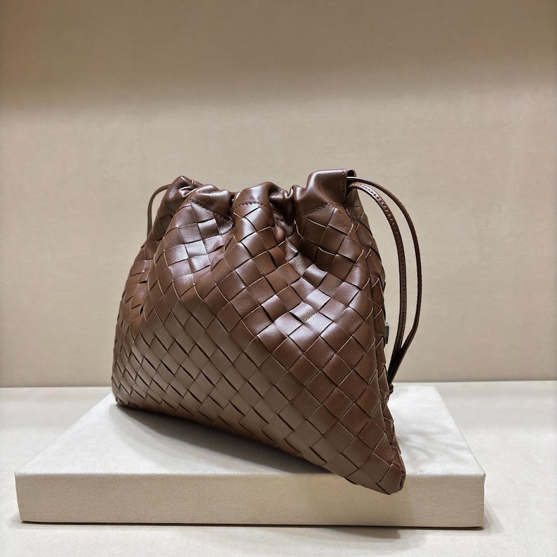 May include: A brown leather handbag featuring a woven intrecciato pattern throughout the exterior. The bag has a soft, slouchy structure with a drawstring closure at the top, secured by thin leather cords that extend from the sides of the opening.