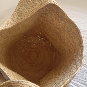 May include: Open view of a woven straw tote bag. The bag's interior is visible, showcasing a circular base and textured walls. The natural beige color of the straw is prominent, with a woven pattern creating a rustic aesthetic. The bag's opening is at the top.