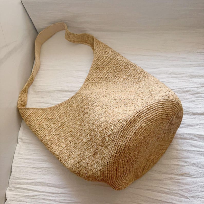 May include: A beige woven shoulder bag with a long strap. The bag has a rounded shape and is made of a textured, natural-looking material. The strap is a lighter shade of beige.